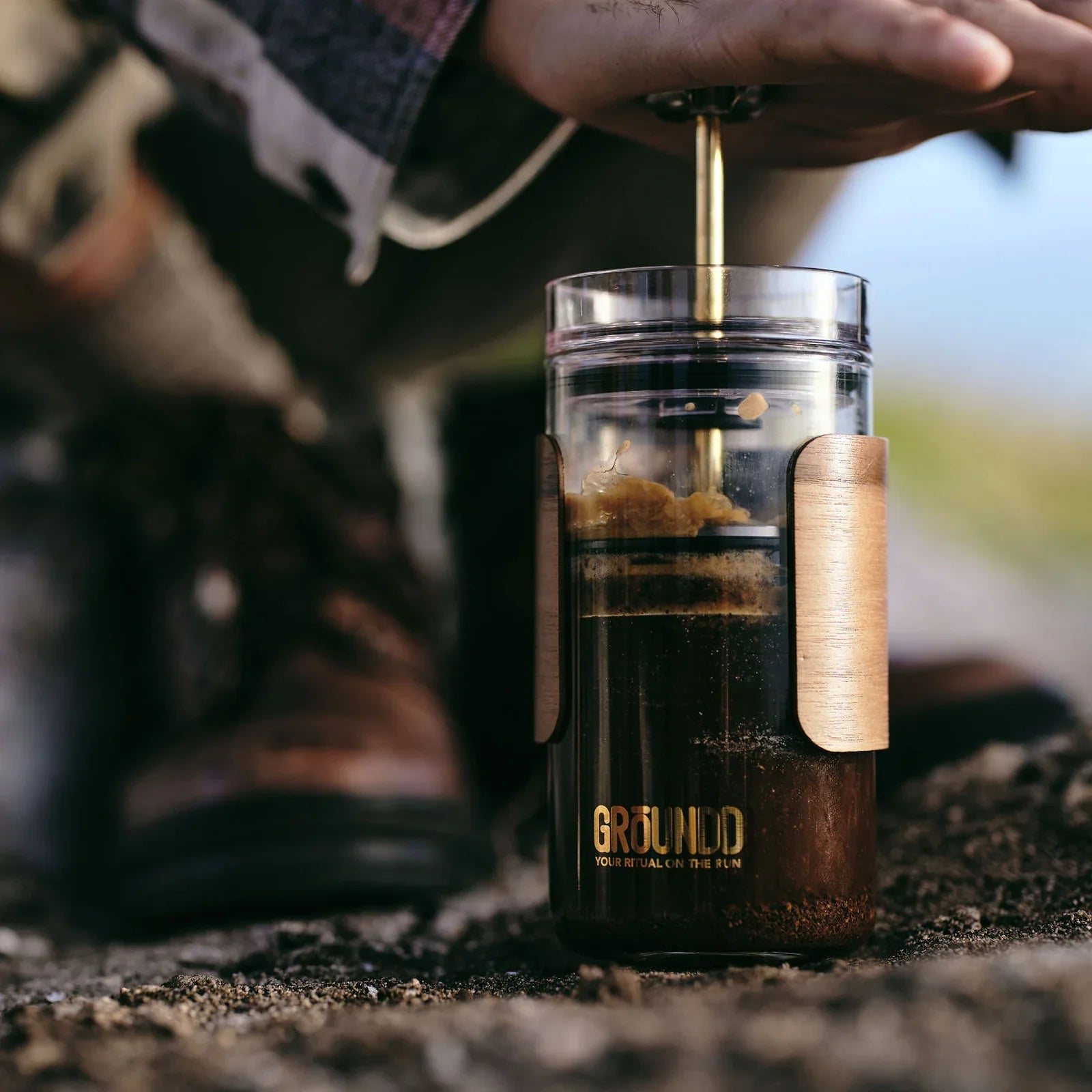 Hand pressing the GROUNDD xpress plunger, coffee brewed on a coastal rock